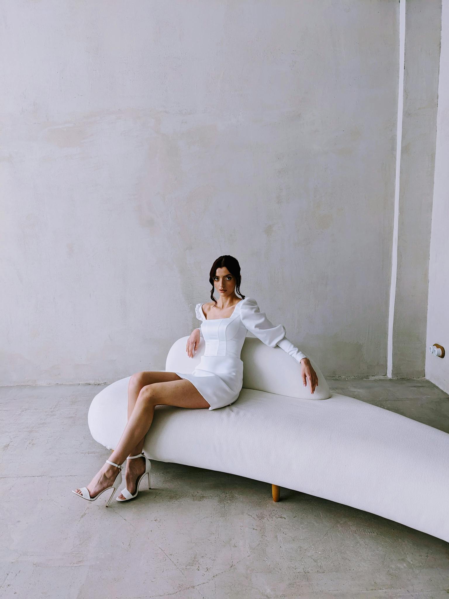 Stylish woman in a white dress elegantly posed on a modern couch in a minimalist setting.